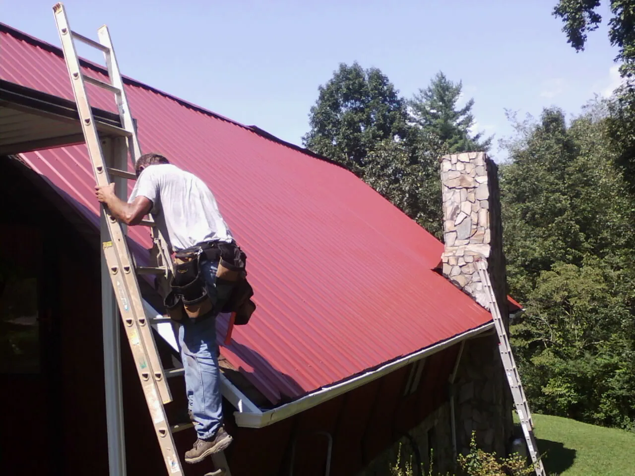 Roofer on a ladder beside a red metal roof and stone chimney — Soffit & Fascia Repair by Roofing Brothers in Middleville